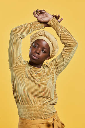 Vertical portrait of confident African-American woman wearing head wrap while dancing against yellow background in studioの写真素材