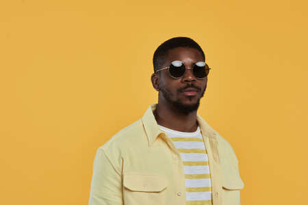 Minimal waist up portrait of trendy African-American man wearing sunglasses and looking at camera while standing against yellow background in studio, copy spaceの写真素材