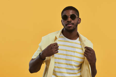 Waist up portrait of handsome African-American man wearing sunglasses and looking at camera with confidence while posing against yellow background in studio, copy spaceの写真素材