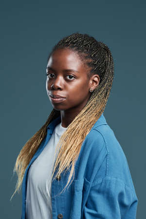 Vertical portrait of modern African-American young woman with Afro braids hairstyle looking at camera while posing against blue backgroundの写真素材