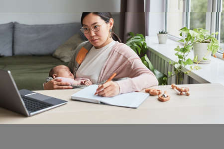 Young woman studying at home, she sitting at the table looking at laptop and making notes in...