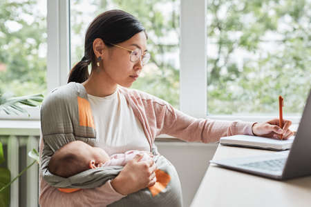 Asian young woman holding her baby on hands while making notes at the table she working at homeの写真素材