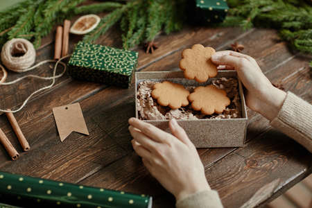 Close up of unrecognizable young woman packing homemade cookies in Christmas gift wrap at rustic wooden table, copy spaceの写真素材