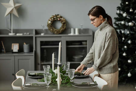 Side view portrait of young woman setting up dining table in dining room decorated for Christmas with fir branches and candles in grey tones, copy spaceの写真素材