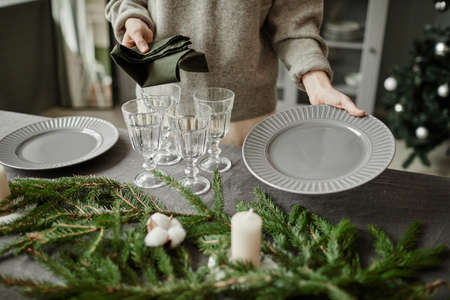 Close up of young woman setting up plates on dining table decorated for Christmas with fir branches and candles in grey tones, copy spaceの写真素材