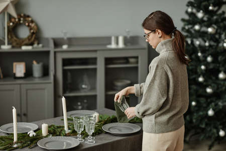 Side view portrait of young woman setting up plates on dining table decorated for Christmas with fir branches and candles in grey tones, copy spaceの写真素材