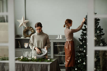 Portrait of two young women setting up dining room decorated for Christmas with fir branches and candles in grey tones, copy spaceの写真素材