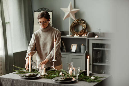 Portrait of young woman setting up dining table decorated for Christmas with fir branches and candles in grey tones, copy spaceの写真素材