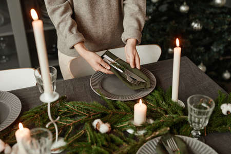 Closeup of unrecognizable woman setting up dining table for Christmas decorated with fir branches and candles in grey tones, copy spaceの写真素材