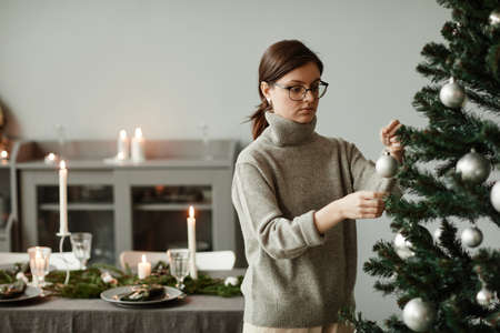 Waist up portrait of young woman decorating Christmas tree at home in elegant silver and grey tones, copy spaceの写真素材