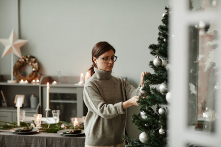Waist up portrait of young woman hanging ornaments on Christmas tree at home in elegant silver and grey tones, copy spaceの写真素材