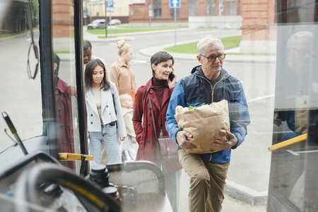 Through glass view at diverse group of people standing in line at bus stop and entering bus