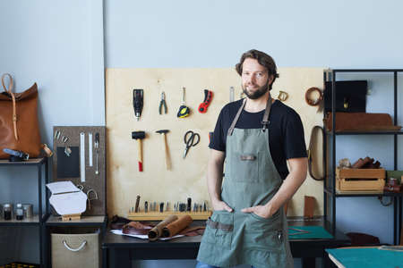 Waist up portrait of smiling male artisan wearing apron and looking at camera while posing in...
