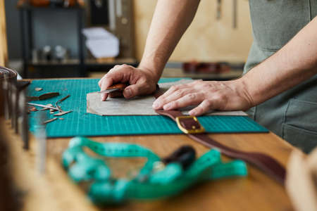 Side view closeup of unrecognizable male artisan making leather belt in tanners workshop,...