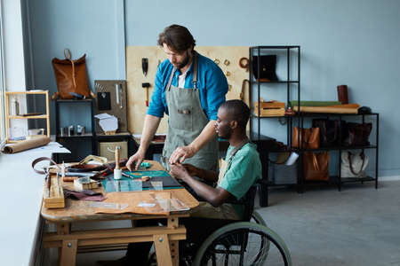 Side view portrait of young African-American man in wheelchair learning leathermaking craft in...