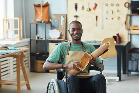 Portrait of smiling African-American man using wheelchair enjoying work in leatherworkers workshop and looking at camera, copy spaceの写真素材
