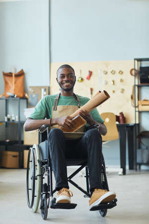 Vertical full length portrait of smiling African-American man using wheelchair enjoying work in leatherworkers workshop and looking at cameraの写真素材