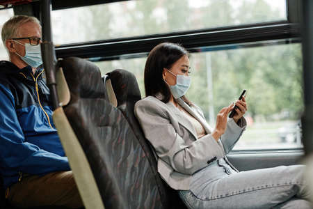 Side view portrait of young Asian woman wearing mask in bus and using smartphone while commuting in city, copy spaceの写真素材