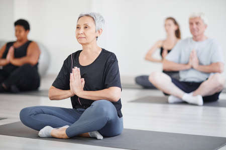 Healthy mature woman sitting on exercise mat in lotus position and doing yoga in the classの写真素材