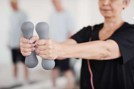 Close-up of senior woman stretching her arms with dumbbells while doing exercises in the gymの写真素材