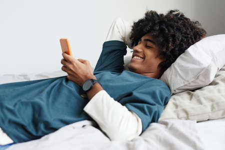 Side view portrait of young African-American man lying on bed at home and smiling while using smartphoneの写真素材