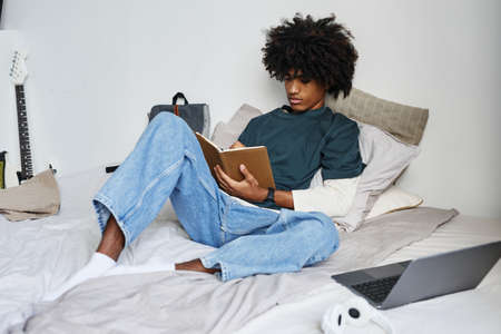Full length portrait of young African-American man sitting on bed at home and writing in notebook, copy spaceの写真素材