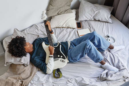 Full length above view at African-American teenager playing guitar while laying on bed at homeの写真素材