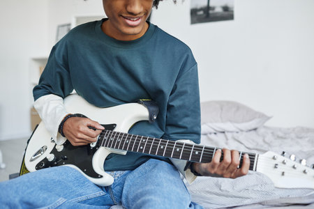 Cropped portrait of smiling African-American teenager playing guitar while sitting on bed at home, copy spaceの写真素材