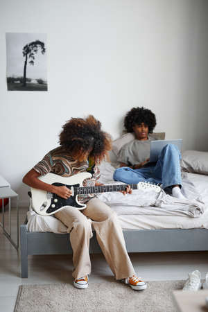 Vertical full length portrait of two teenagers at home, focus on young African-American woman playing electric guitarの写真素材