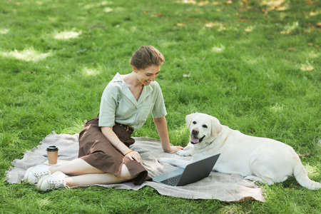 Full length portrait of happy young woman enjoying picnic in park with white Labrador dog and using...