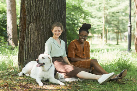 Full length portrait of two young women sitting on green grass in park with Labrador dog and...
