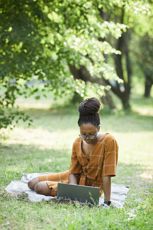 Vertical portrait of African-American young woman using laptop in park while sitting on grass, copy spaceの写真素材