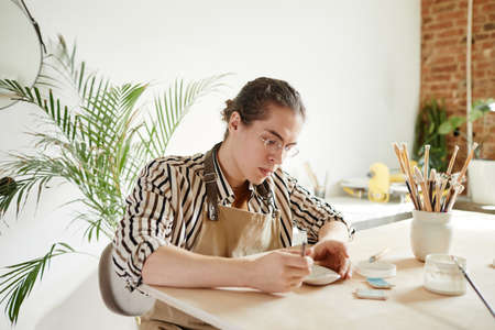 Warm toned portrait of young male artist decorating ceramics in pottery workshop, copy spaceの写真素材