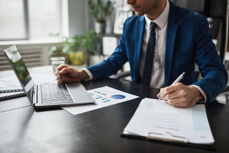 Cropped portrait of successful businessman in suit working at able in office and using laptop, copy...