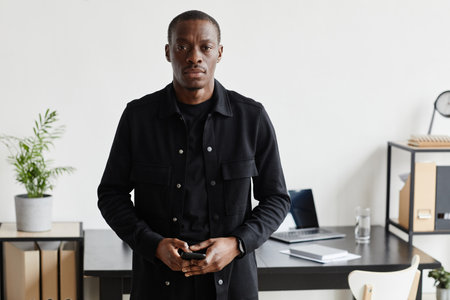 Waist up portrait of African-American businessman looking at camera while standing by desk at...