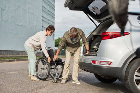 Full length portrait of mature couple unloading wheelchair out of car trunk in parking lot outdoors, copy spaceの写真素材