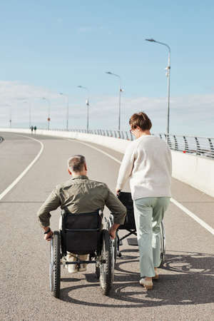 Vertical back view portrait of mature couple with man in wheelchair walking on road at bridge, copy spaceの写真素材