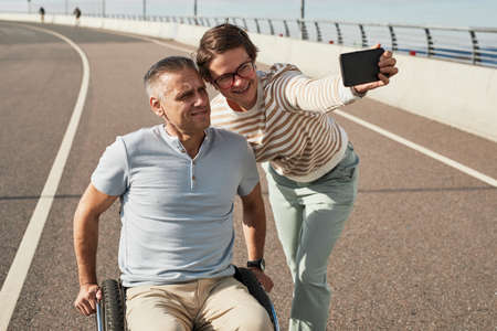 Portrait of adult couple with man in wheelchair taking selfie outdoors and enjoying city life, copy spaceの写真素材