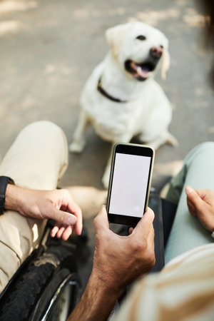 Close up of adult man in wheelchair using smartphone with blank screen, dog in background, copy...