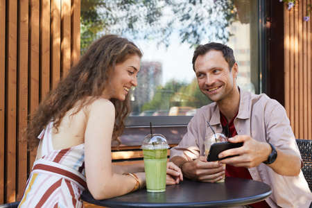 Portrait of happy couple looking at smartphone screen at outdoor cafe while enjoying date in Summer,...