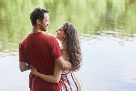 Back view portrait of happy young couple embracing while enjoying romantic date outdoors by water, copy spaceの写真素材