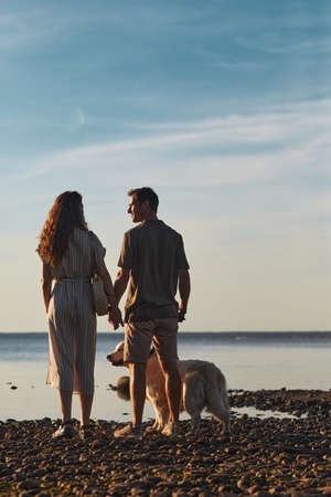 Back lit full length portrait of happy couple with dog together by water at sunsetの写真素材