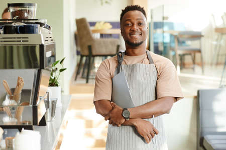 Horizontal medium portrait of joyful African American man wearing apron working in cafe holding pen...
