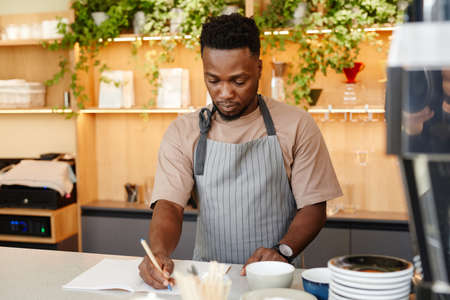 Horizontal medium portrait of young Black man wearing apron standing at cafe kitchen counter writing something in notebookの写真素材