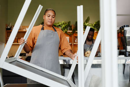Young adult African American man and woman starting workday in modern cafe preparing tables and...