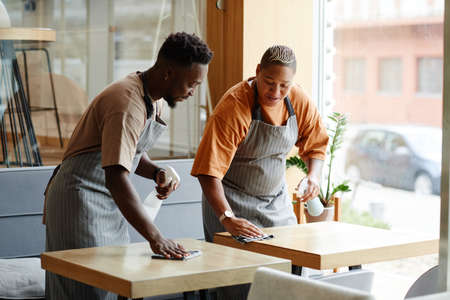 Young African American man and woman wearing aprons working in small cafe preparing tables for customers cleaning them with spray detergent and chattingの写真素材