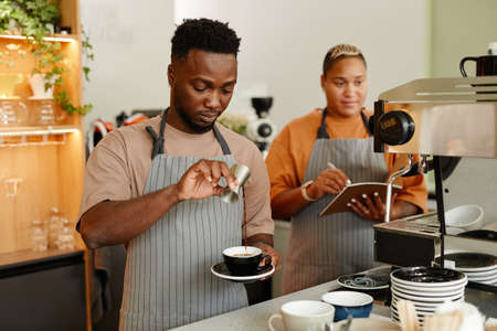 Young African American man and woman wearing aprons working in small cafe making coffee drink and writing something in notebookの写真素材