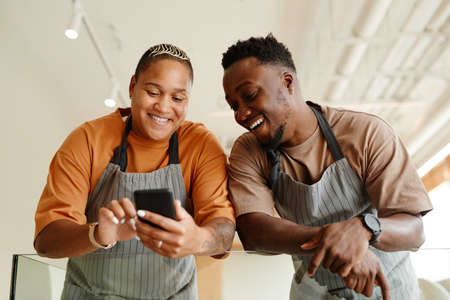 Horizontal low angle shot of young African American man and woman working together in modern cafe watching something in Internet during breakの写真素材