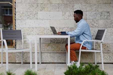 Side view shot of young African American man wearing casual outfit sitting at cafe table outdoors using laptopの写真素材