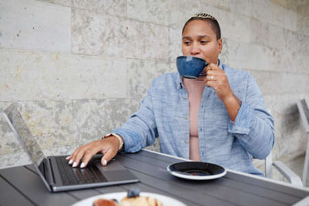 Portrait of young African American woman wearing casual outfit sitting at cafe table outdoors using...